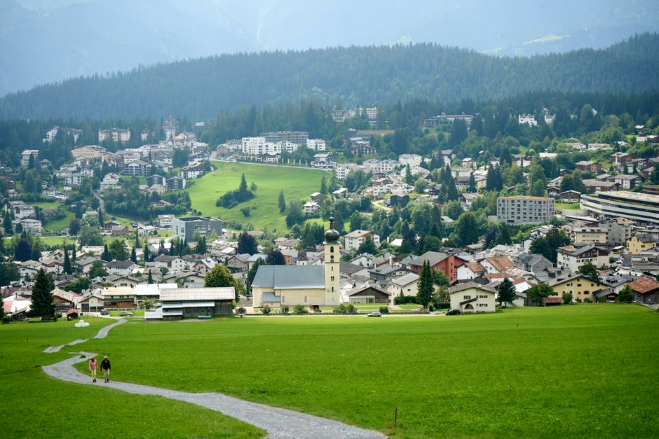 Aerial view of a charming Alpine town nestled in lush green hills under a clear summer sky.