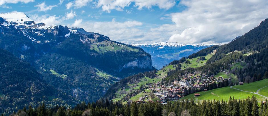 Picturesque scenery of small village located on green hill surrounded by mountains covered by green forest in sunny day
